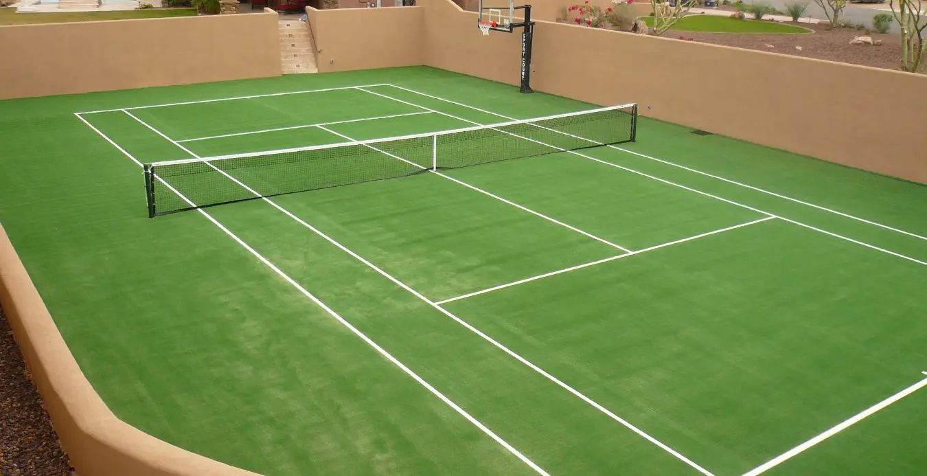 An outdoor court in Queen Creek AZ with green artificial lawn and white lines, set up for a tennis game. It features a tennis net in the center and a basketball hoop on one end. The court is enclosed by a tan wall, with steps leading up from the court level. Surrounding the area is desert landscaping.