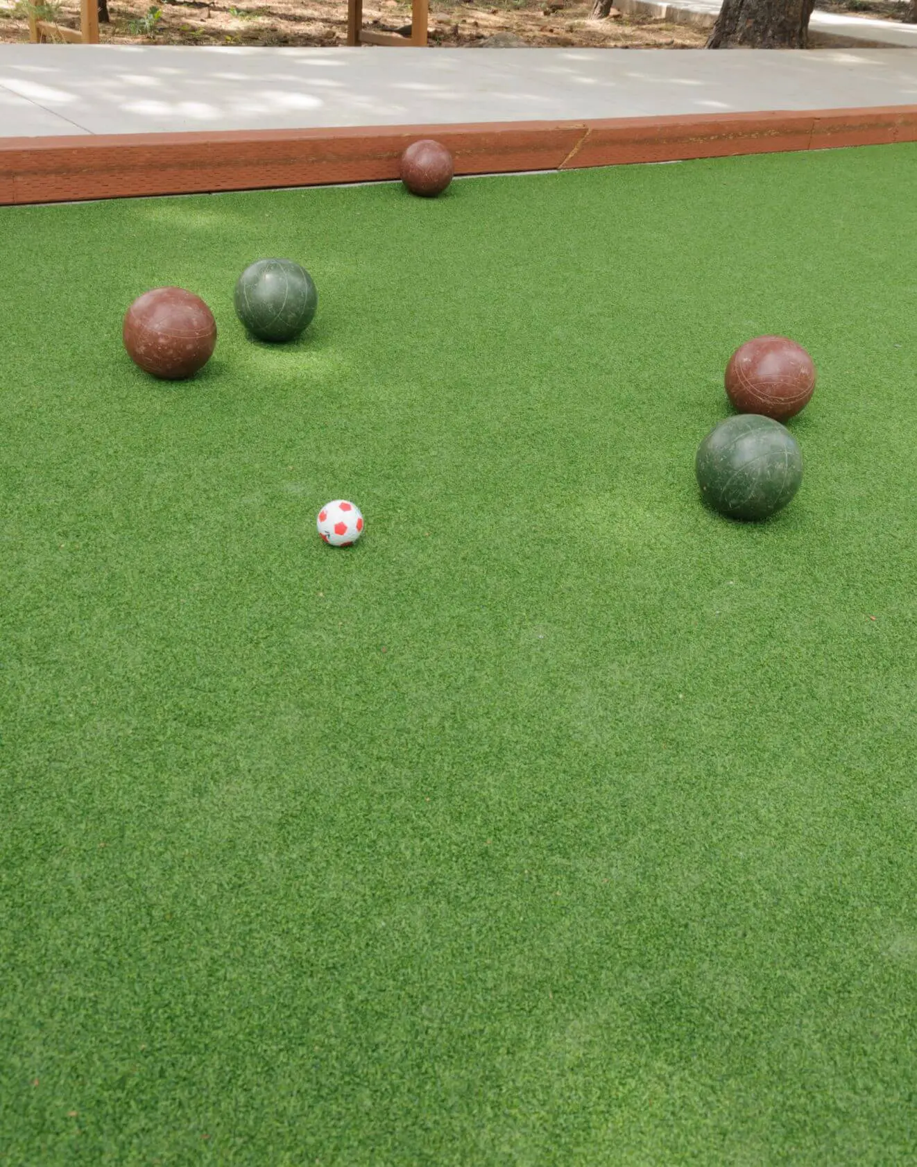 A bocce ball court with green artificial turf in Queen Creek, AZ. Five balls are visible: three larger balls (two brown and one green) near the top half of the court, and one red and white polka-dotted smaller ball closer to the bottom. The court, expertly crafted by athletic turf installation specialists, is bordered with wooden edges.