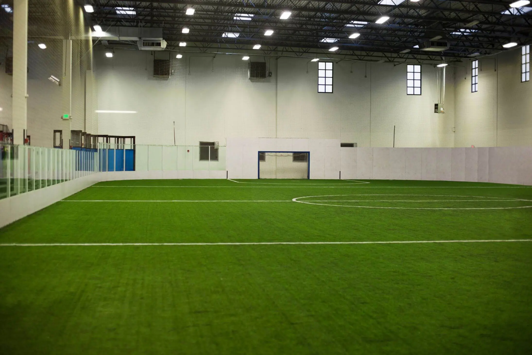 Indoor soccer field with green artificial turf, white walls, and clear barriers. The ceiling is high with visible lighting fixtures, and there are several windows along the back wall. Featuring professional athletic turf installation, the field has white boundary lines and a goal at the far end.