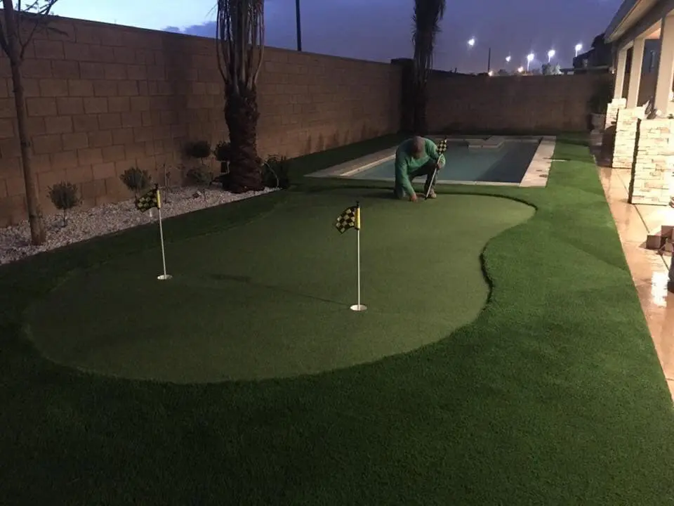 A person is kneeling on an artificial grass putting green in a backyard in Queen Creek, AZ. The meticulously designed green features two holes, each marked with a small flag. Surrounded by manicured artificial turf and adjacent to a sleek rectangular swimming pool and tall boundary wall, the scene is immaculate.