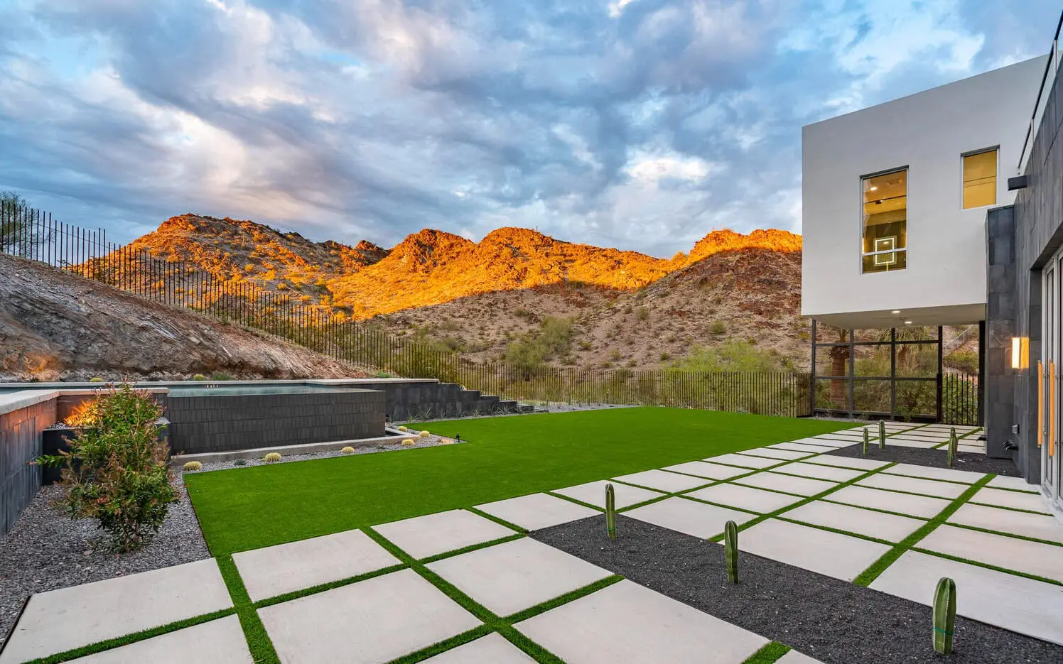 A modern house with large windows and a minimalist backyard featuring a green lawn, concrete pavers, and small cacti. The background showcases rugged mountains bathed in golden sunlight under a partly cloudy sky, accentuating the appeal of the putting greens created by artificial turf installation.