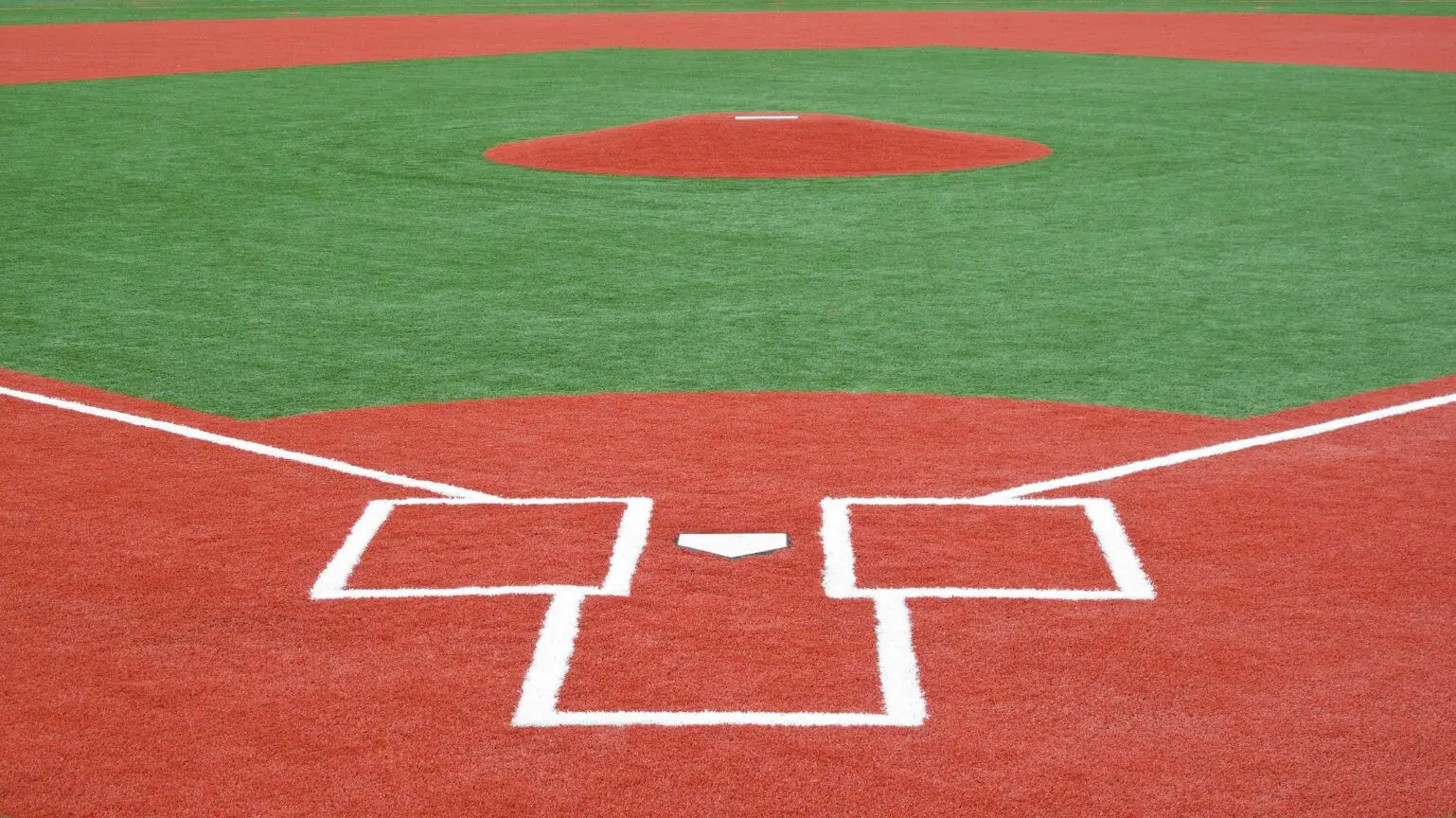 A baseball field in Queen Creek, AZ with a view from home plate, featuring the pitcher's mound in the distance. The field showcases an impressive athletic turf installation with clear demarcations of the batter's box, home plate, and pitcher's mound. The red and green artificial lawn contrasts sharply.