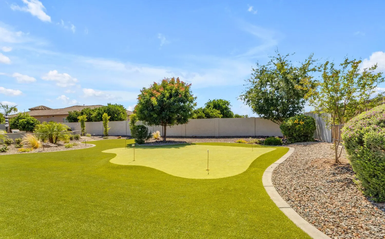 A well-maintained backyard putting green, featuring athletic turf installation, is surrounded by trimmed bushes and trees under a clear blue sky in Queen Creek, AZ. The area is bordered by gravel and a concrete edge, with two golf flags planted in the green. A house and fence can be seen in the background.