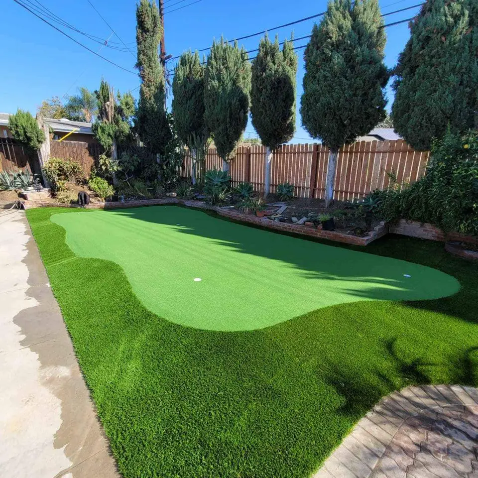 A backyard scene featuring a small putting green, surrounded by neatly trimmed artificial grass. Tall trees and various plants line the wooden fence enclosing the area. The sky is clear and blue, enhancing the serene and tidy look of the space, showcasing professional artificial turf installation.