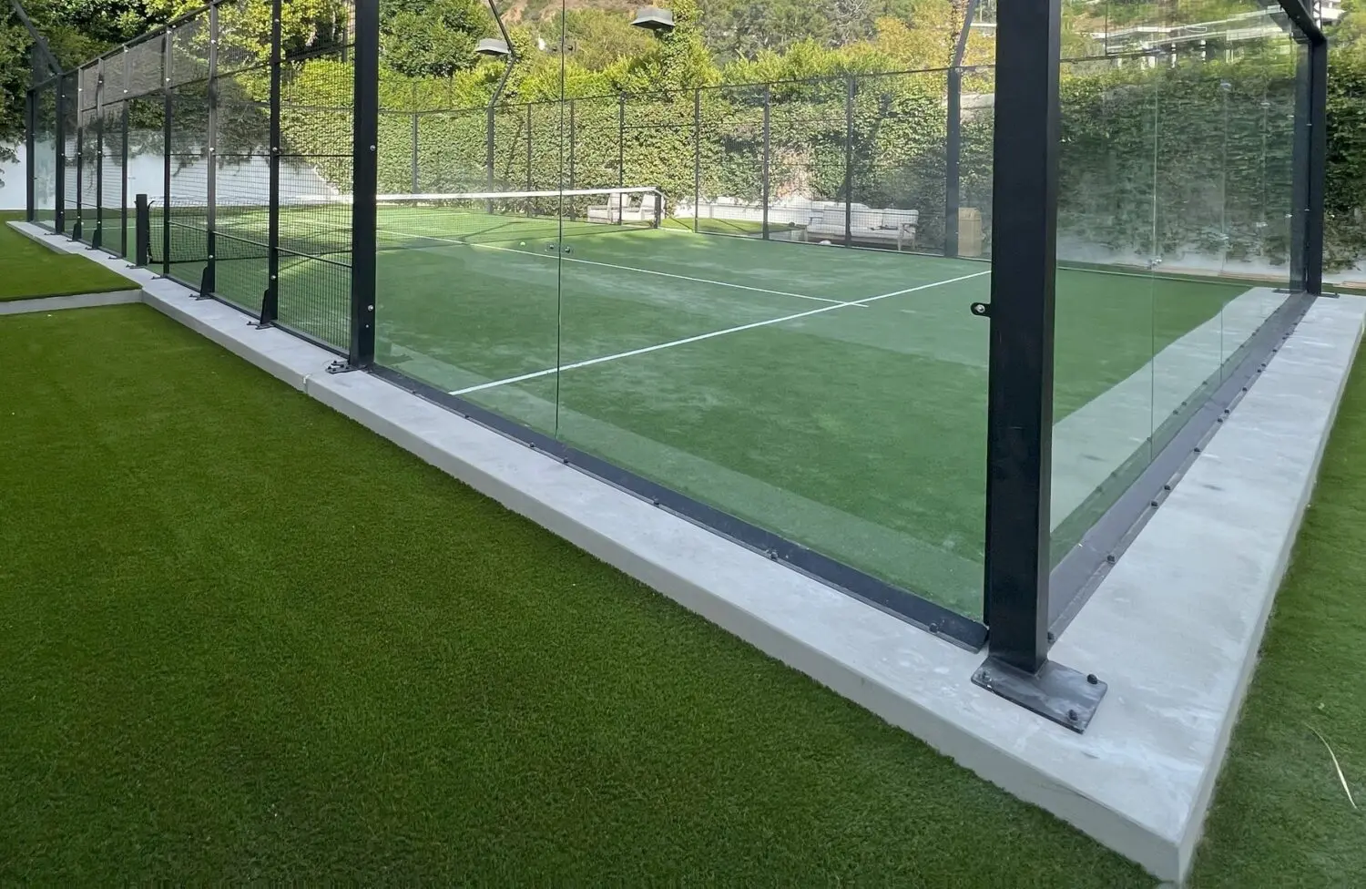 A padel court with glass walls and netting, enclosed by a fence on a foundation of athletic turf installation. The background features dense greenery, including trees and bushes, creating a serene outdoor setting in Queen Creek AZ.