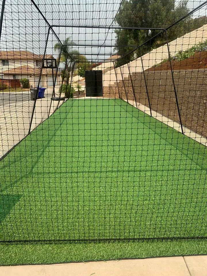 A rectangular outdoor batting cage with a green synthetic grass floor, enclosed with black netting. The cage is situated in a residential backyard with a sloped retaining wall on the right and a basketball hoop visible in the background, seamlessly blending into the Goodyear landscape.