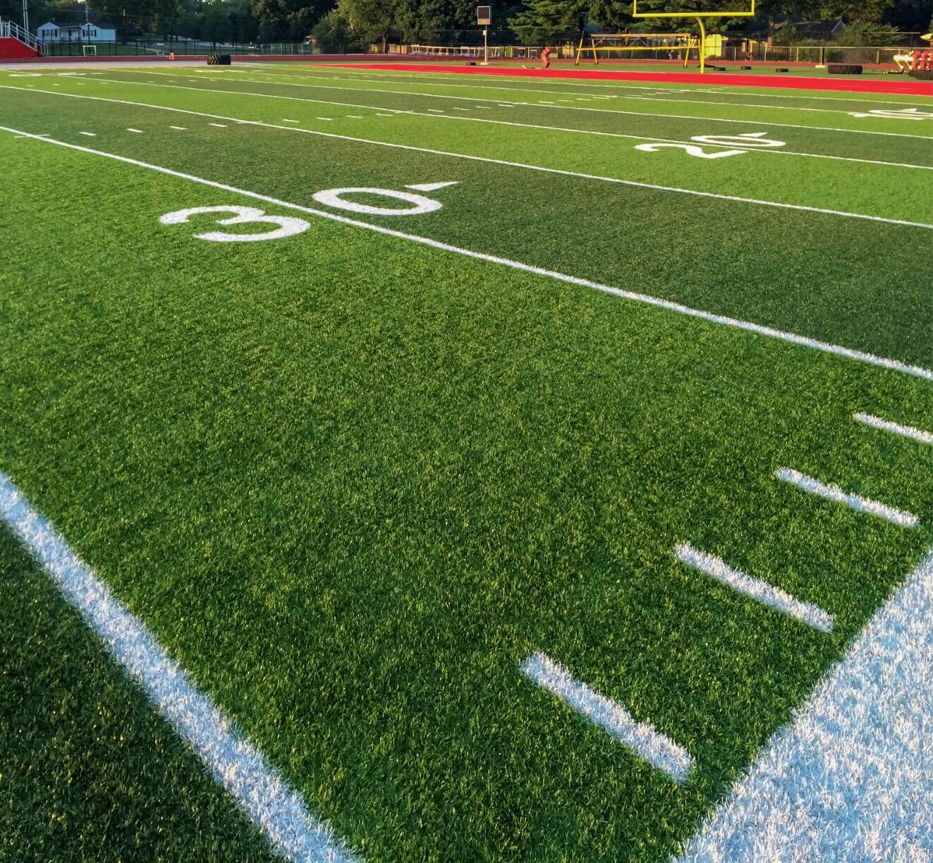 Close-up of a green football field with white yard markers, showcasing the 30-yard line prominently. The athletic turf installation extends towards a yellow goalpost in the distance. Bleachers can be seen in the far background under a blue sky.