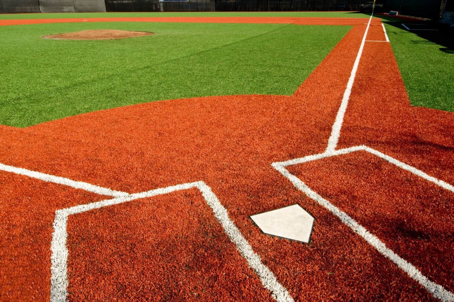 Close-up view of a baseball diamond in Queen Creek AZ featuring home plate in the foreground, surrounded by a red artificial turf surface with white foul lines. The pitcher's mound is visible in the distance, with a contrasting green field thanks to top-notch athletic turf installation.