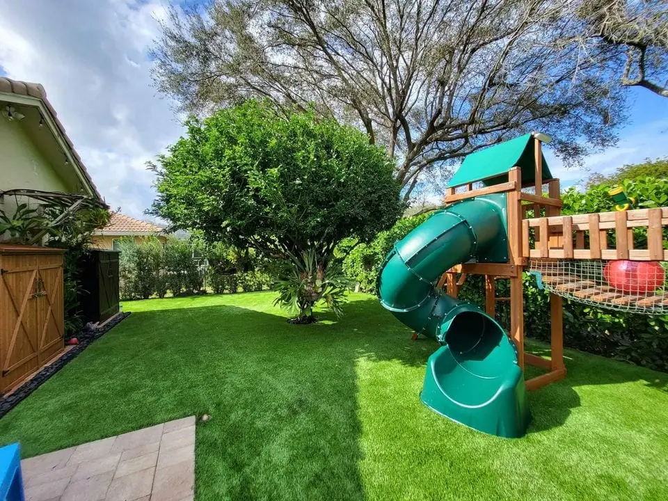 A backyard in Queen Creek featuring a playset with a green tube slide, wooden climbing structure, and small playhouse. The yard boasts lush artificial grass installation, a large leafy tree, and a wooden fence on the left. The sky is partially cloudy, and the area is well-lit by sunlight.