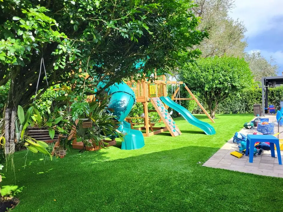 A backyard playground in Queen Creek features a wooden play structure with a blue slide and a spiral tube slide, surrounded by greenery. Nearby, a patio with blue chairs and a table holds various toys and items. The area boasts lush, well-manicured grass enhanced by professional play turf installation.