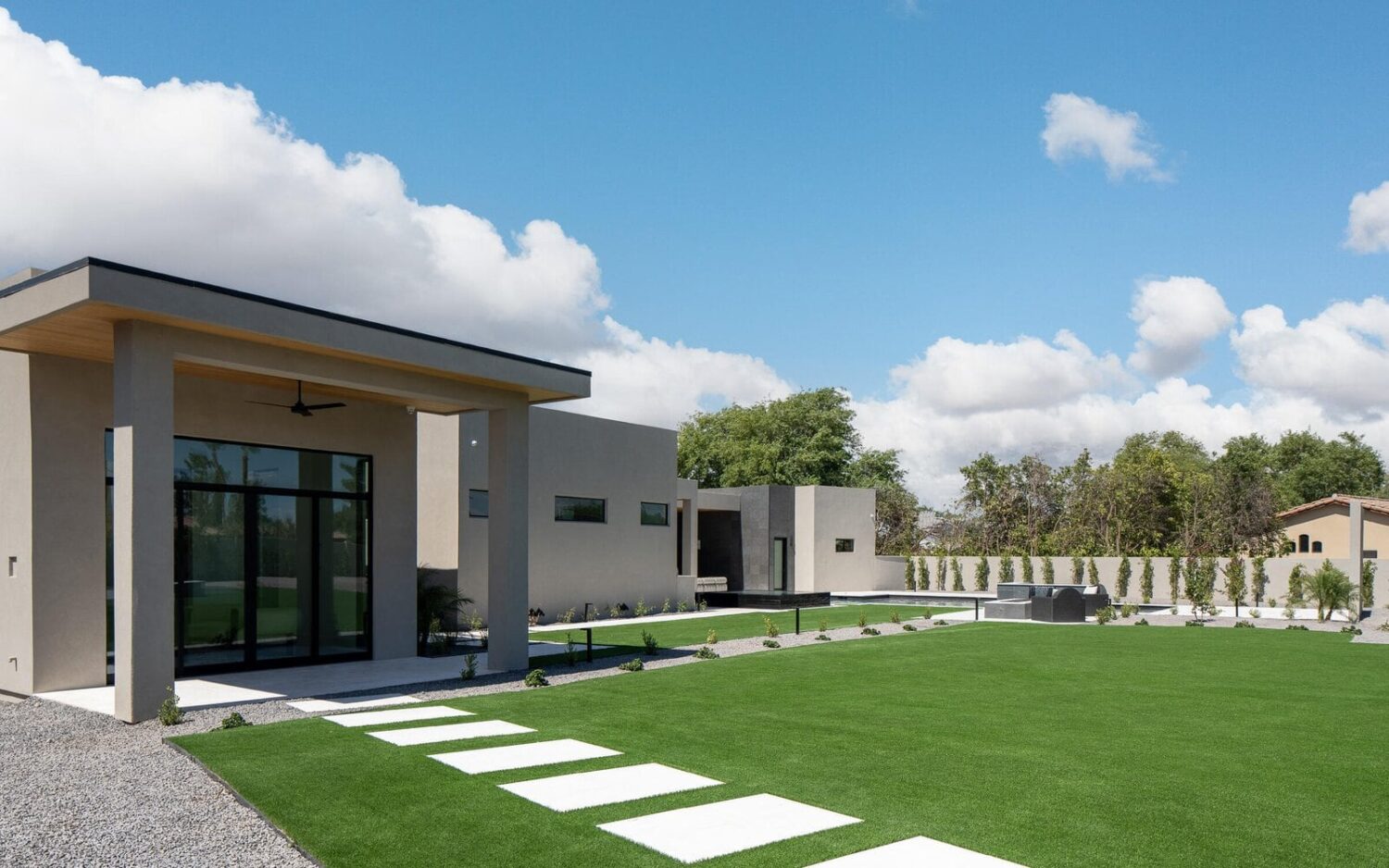 Modern house exterior with minimalist design featuring large glass doors, flat roofs, and clean lines. The Goodyear landscape boasts a well-manicured lawn with stone stepping paths and synthetic grass installations for easy maintenance. The sky is blue with scattered white clouds.