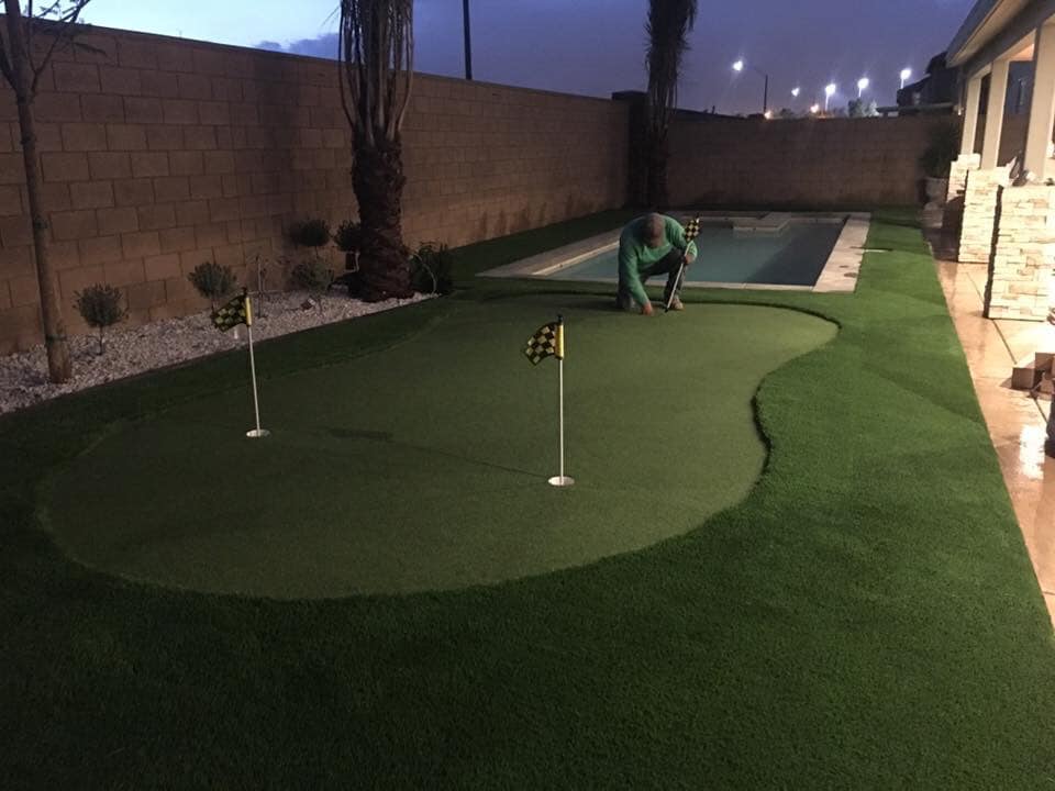 A person kneels on a Goodyear Greens Turf backyard putting green, adjusting a flag. The area includes two holes with checkered flags. A narrow swimming pool is to the right, and the scene is illuminated in the evening light, showcasing a flawless artificial grass installation.