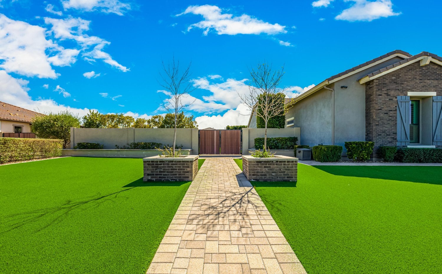 A beautifully landscaped backyard featuring a stone-paved pathway leading to a wooden gate. The path is flanked by lush synthetic grass lawns and two brick planters holding small, leafless trees. Installed by Goodyear Greens Turf, the vibrant greenery contrasts with the bright blue sky and scattered white clouds.